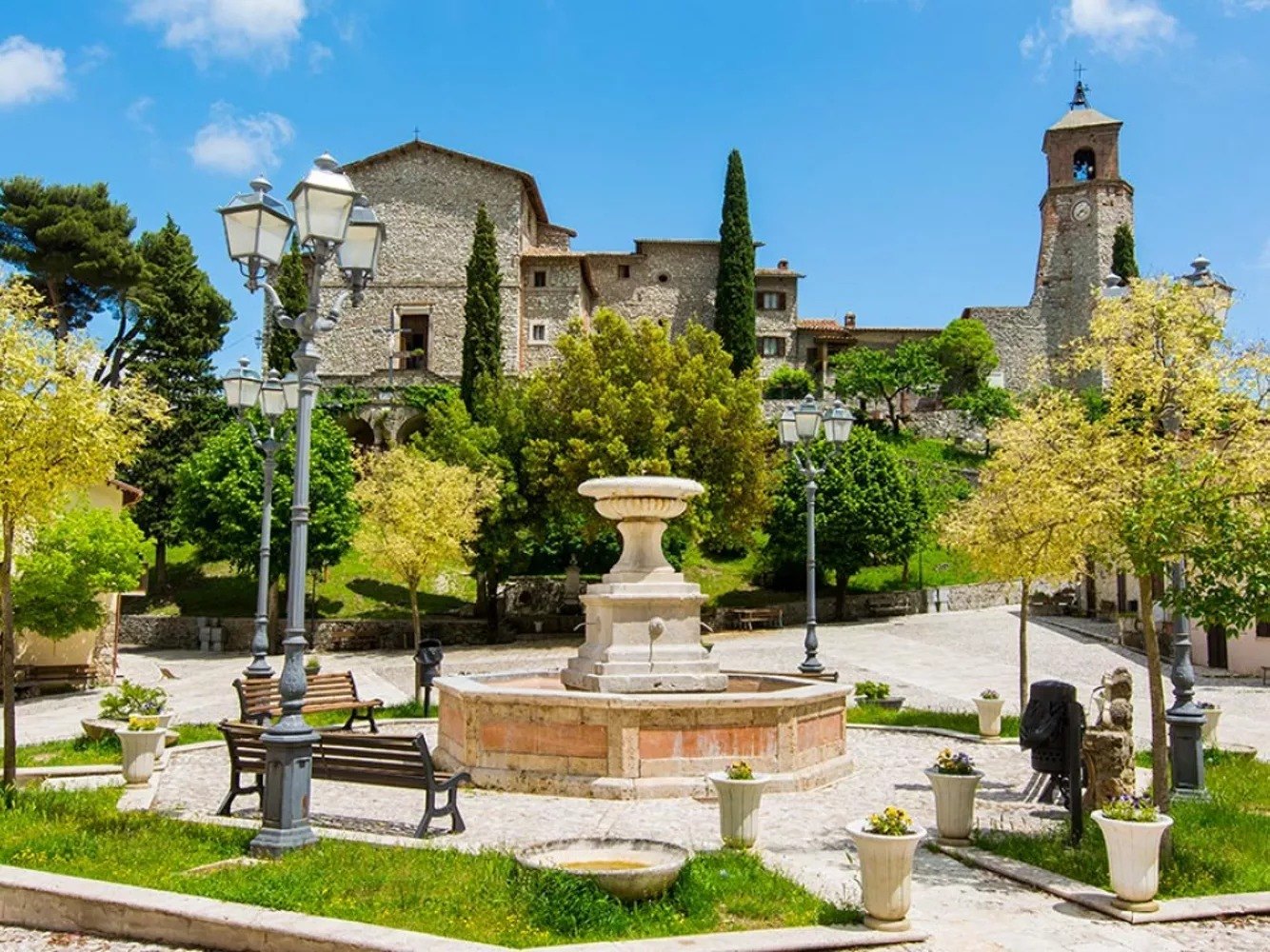 La piazza di Greccio di giorno, con la fontana in travertino, le panchine, i lampioni e la torre dell'orologio